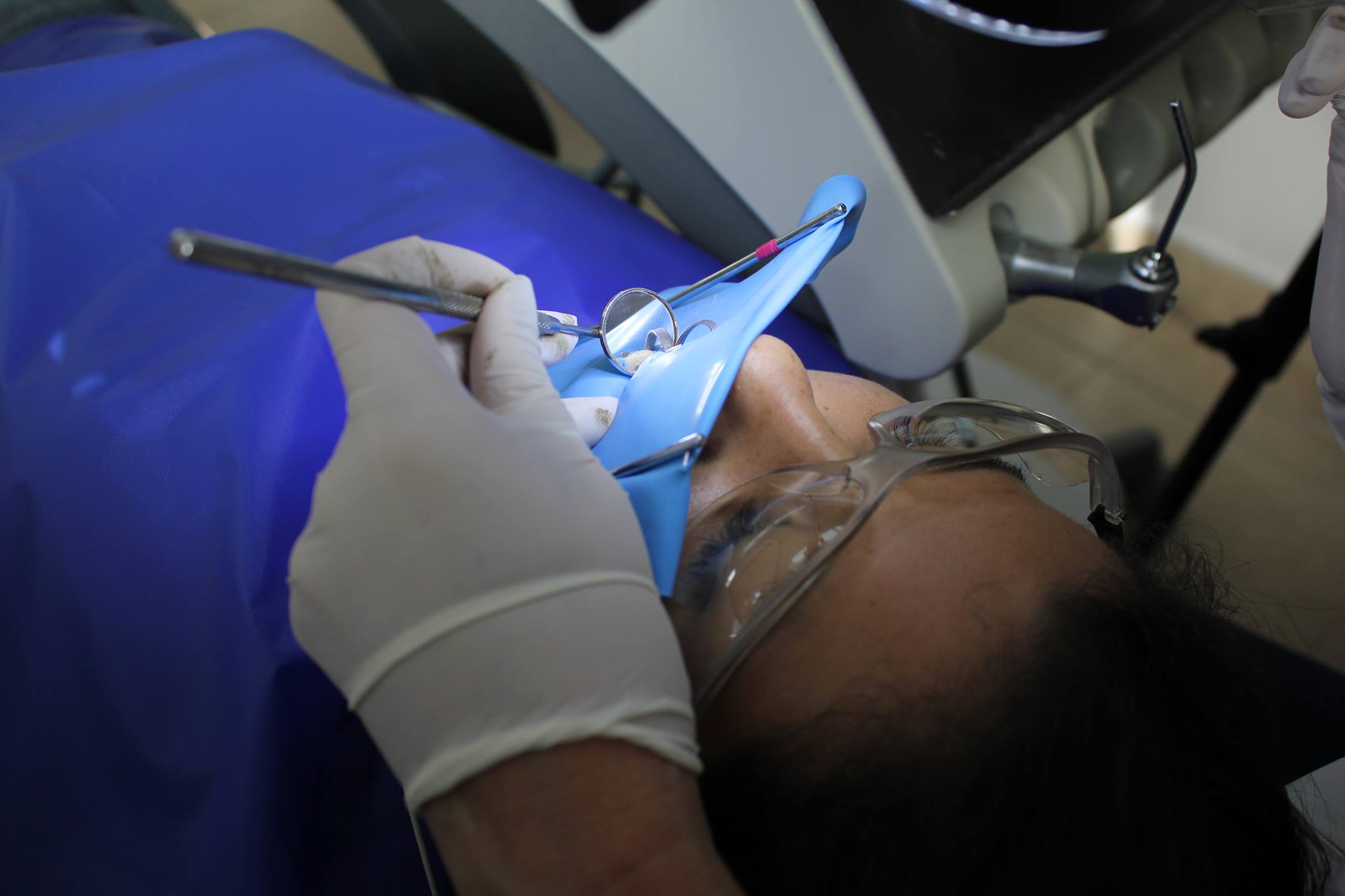 Endodoncia Dentista con guantes examina la boca de un paciente con dique de goma azul y gafas protectoras.