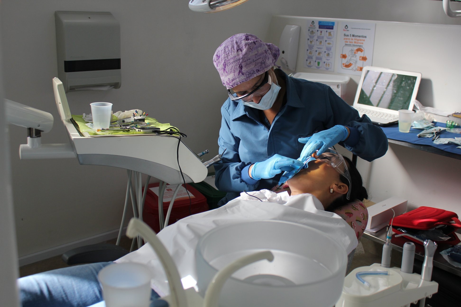 Dentista con mascarilla y guantes azules realizando un procedimiento dental a una paciente reclinada en el sillón.